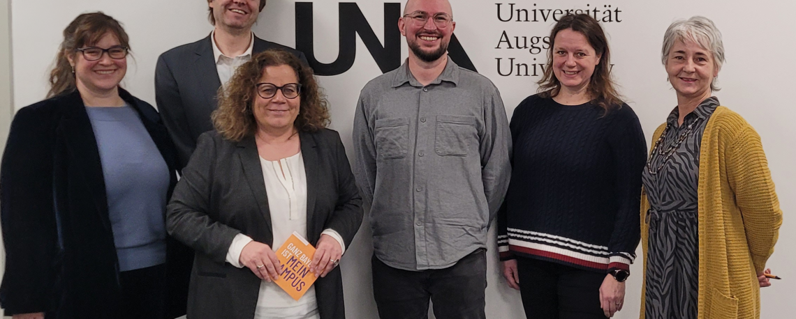 Foto von links: Dr. Katrin Bauer (Instructional Designerin am DigiLLab), Dr. Carl-Christian Fey (Geschäftsführer des DigiLLab), Katja Graßl (Kanzlerin der Uni Augsburg), Prof. Dr. Michael Sailer (wissenschaftlicher Direktor des DigiLLab), Henrike Wackermann-Eckert (Koordinatorin des Erweiterungsfach Medienpädagogik) und Regina Bauer (Sekretariat des DigiLLab) vor dem Logo der Uni Augsburg