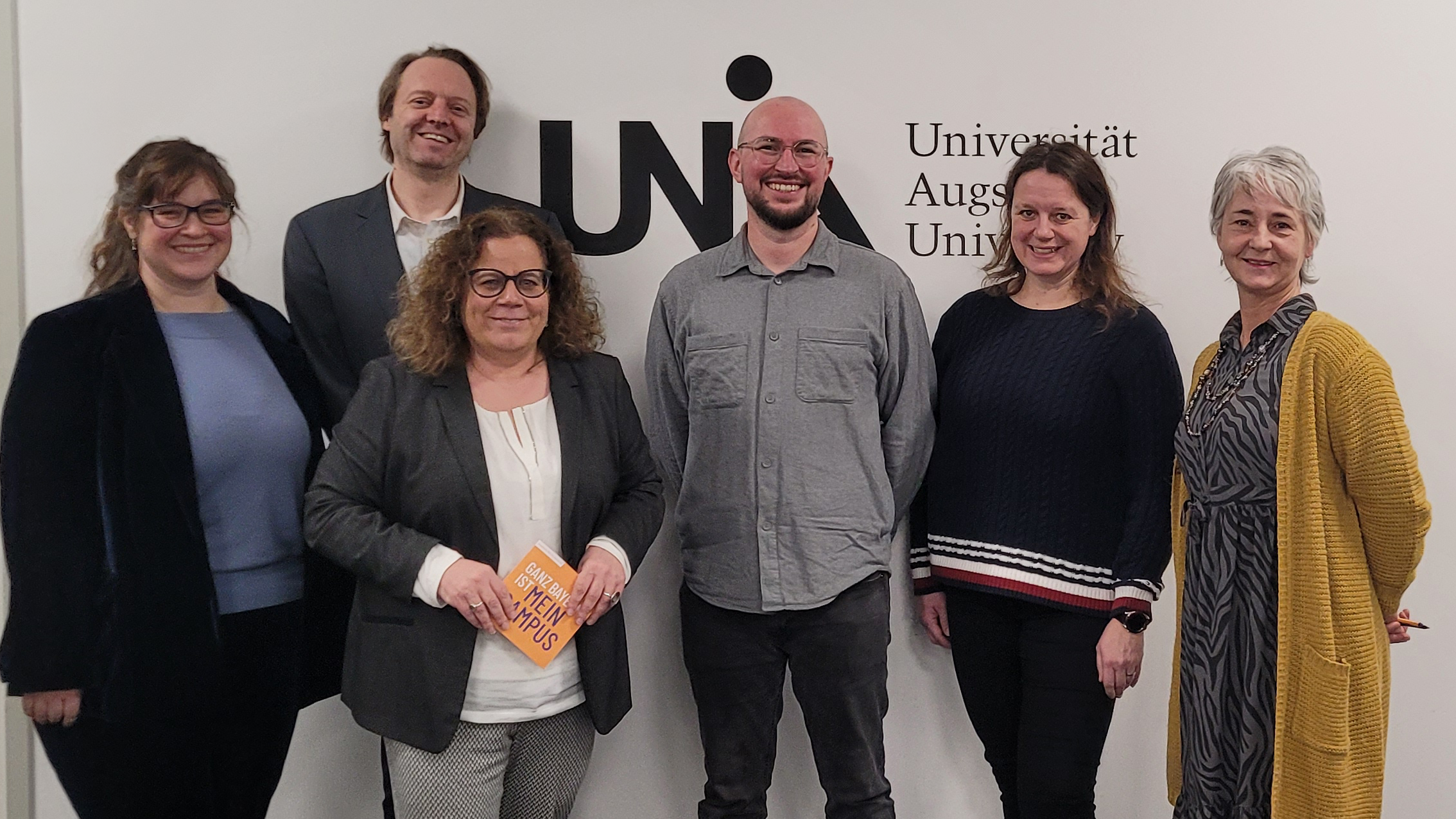 Foto von links: Dr. Katrin Bauer (Instructional Designerin am DigiLLab), Dr. Carl-Christian Fey (Geschäftsführer des DigiLLab), Katja Graßl (Kanzlerin der Uni Augsburg), Prof. Dr. Michael Sailer (wissenschaftlicher Direktor des DigiLLab), Henrike Wackermann-Eckert (Koordinatorin des Erweiterungsfach Medienpädagogik) und Regina Bauer (Sekretariat des DigiLLab) vor dem Logo der Uni Augsburg